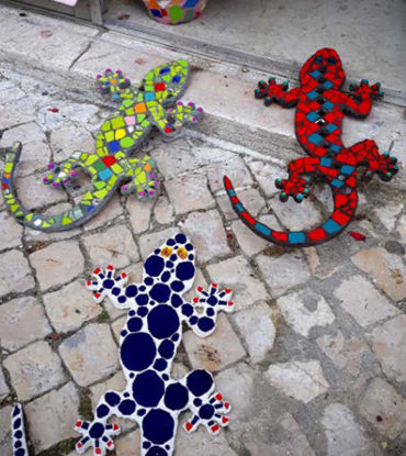 Three colorful mosaic geckos made of ceramic tiles, each with unique designs, arranged on the ground in a vibrant display.